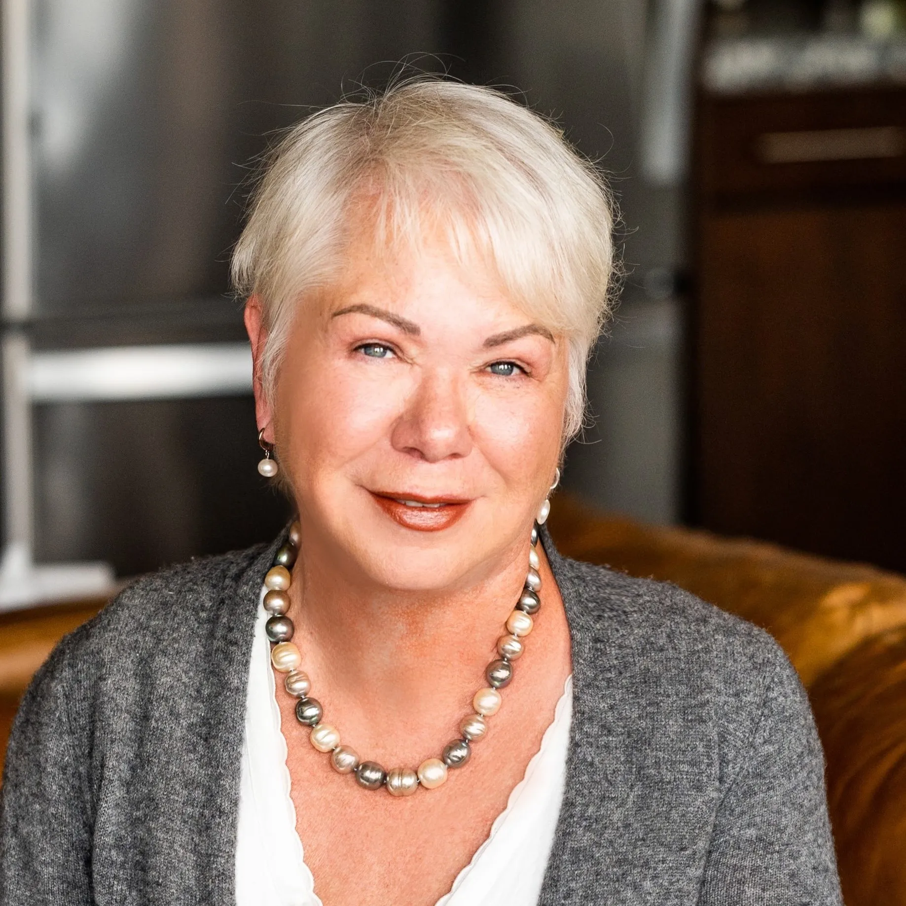 Photograph of a white haired, blue eyed woman wear a beautiful pearl necklace, grey sweater and white dress.