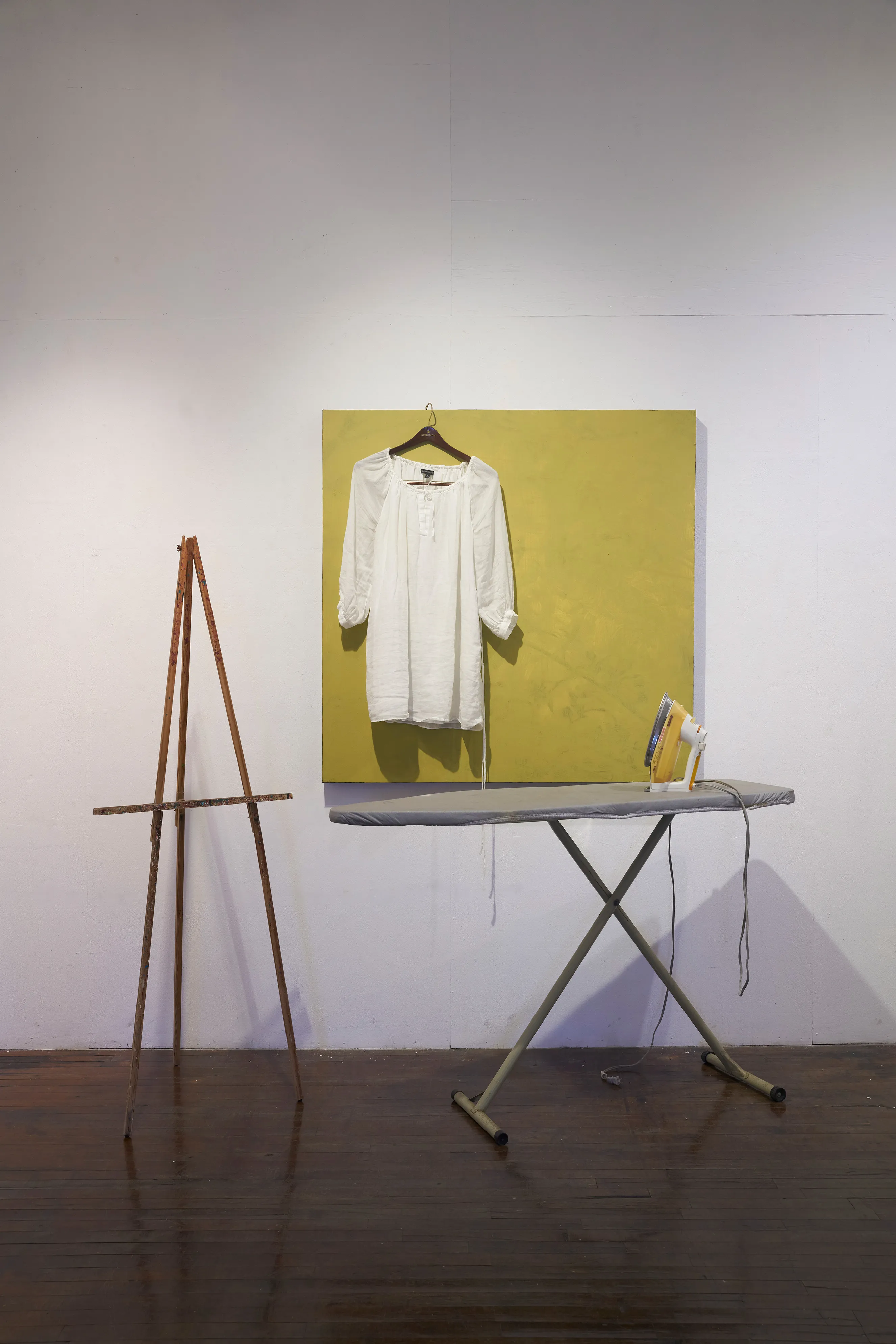 White dress hanging off of a canvas mounted on the wall. To the right of the dress in the foreground is an ironing and iron, and to the left of the dress is an easel.